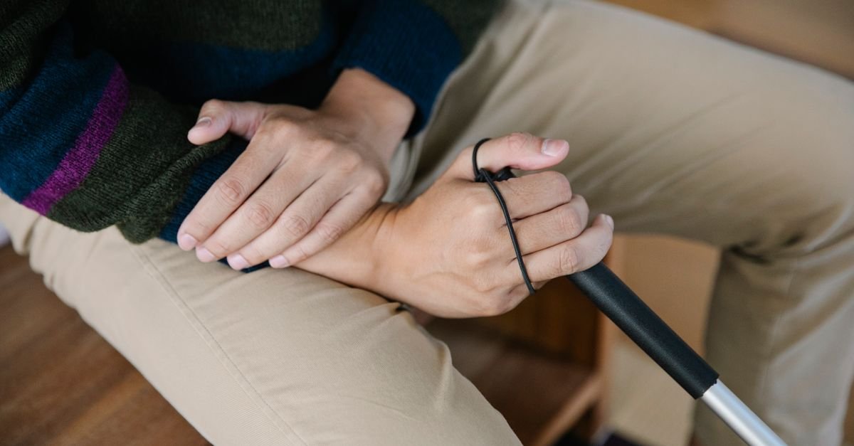 Mãos de pessoa idosa apoiadas em uma bengala, sentada em ambiente interno