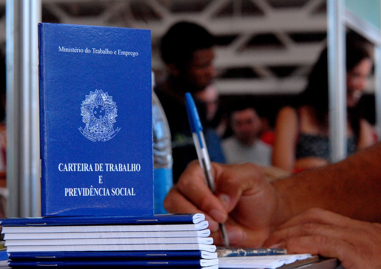 Carteira de Trabalho e Previdência Social em destaque sobre mesa, com pessoa preenchendo formulário ao fundo