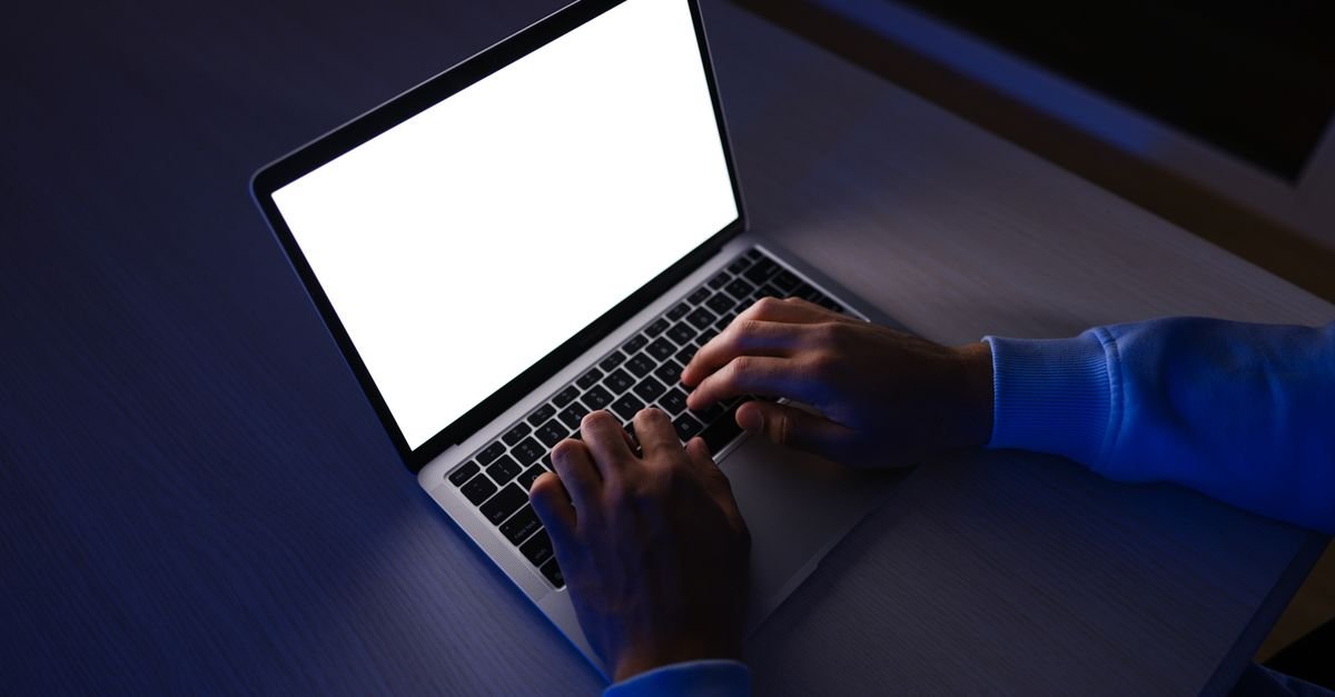 Mãos digitando em um notebook com a tela acesa em um ambiente escuro