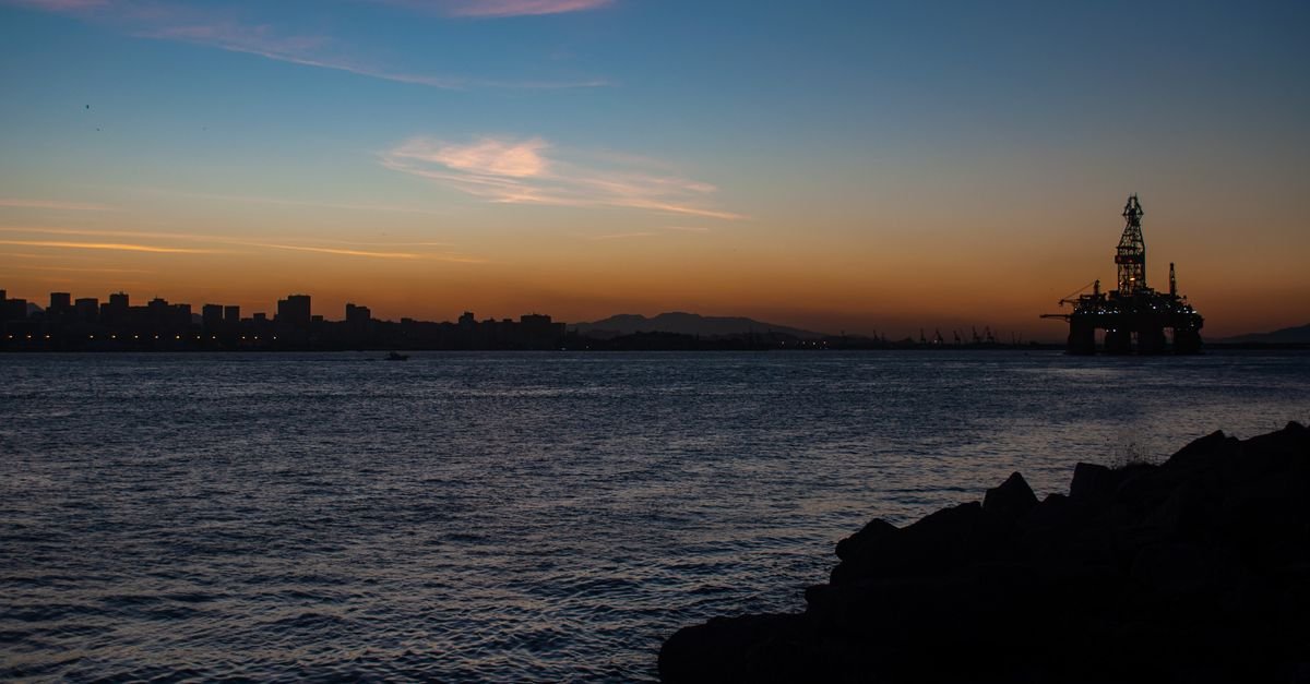 Plataforma de petróleo offshore em silhueta ao entardecer, com a costa ao fundo