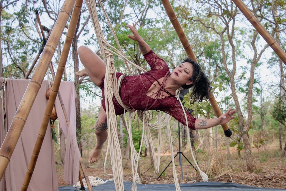 Acrobata de vestido vermelho suspensa em cordas de macramê presas a uma estrutura de bambu