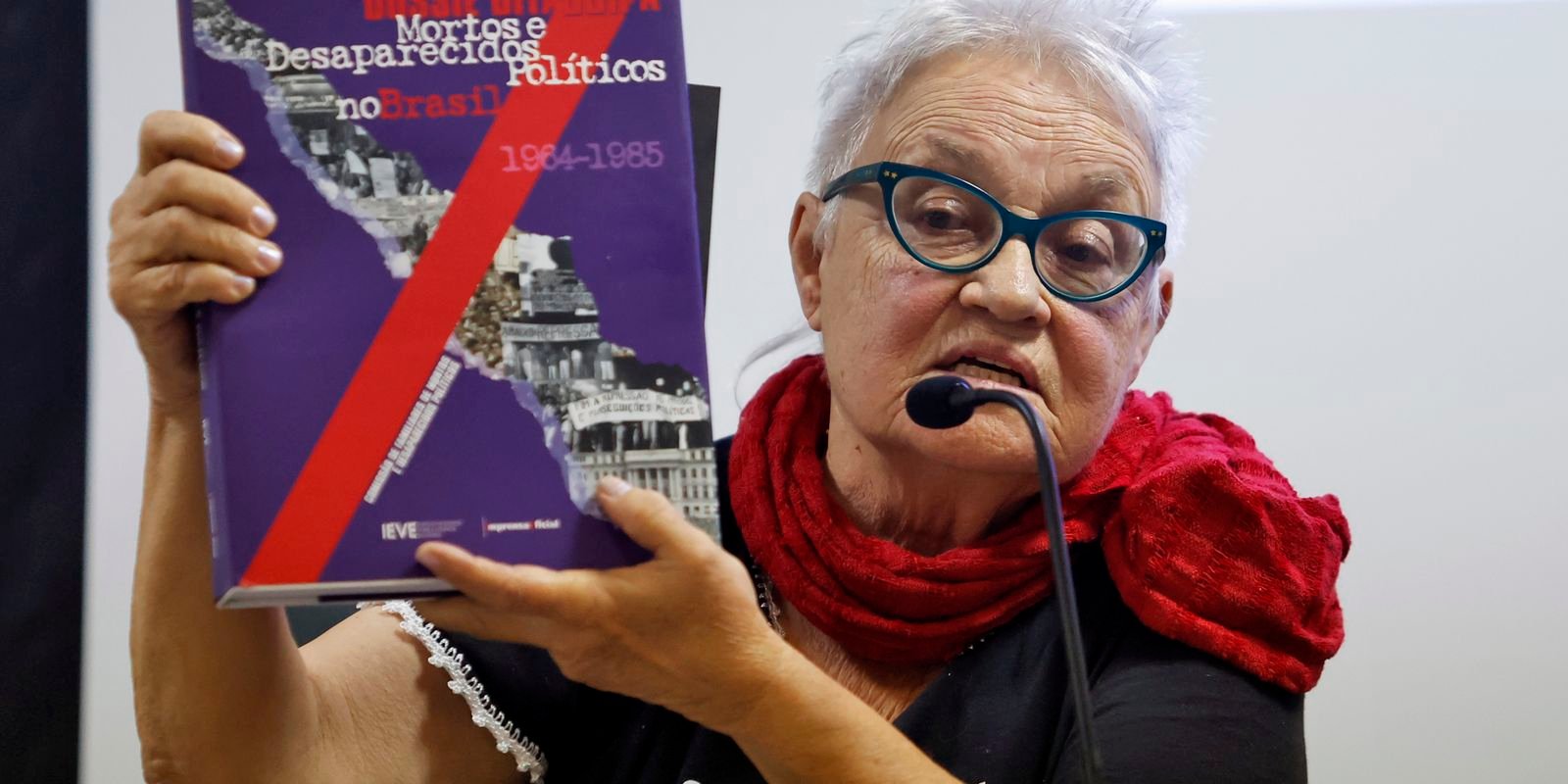 Amelinha Teles segura exemplar do dossiê sobre mortos e desaparecidos políticos enquanto fala ao microfone