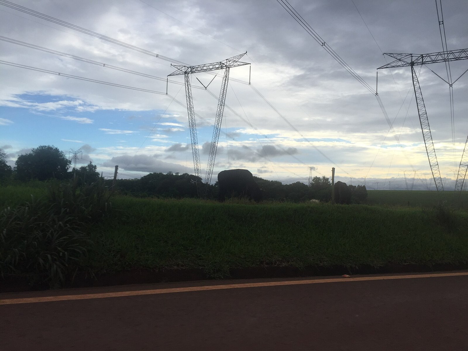 Torres de linha de transmissão de energia elétrica em área rural ao amanhecer, ao lado de rodovia