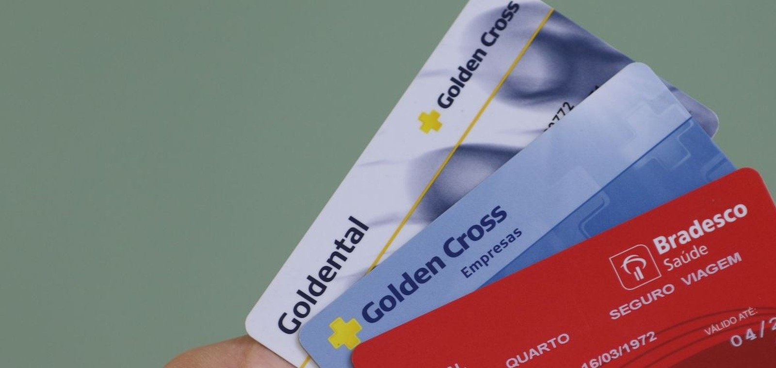 Mão segurando três carteirinhas de planos de saúde: Goldental, Golden Cross e Bradesco Saúde