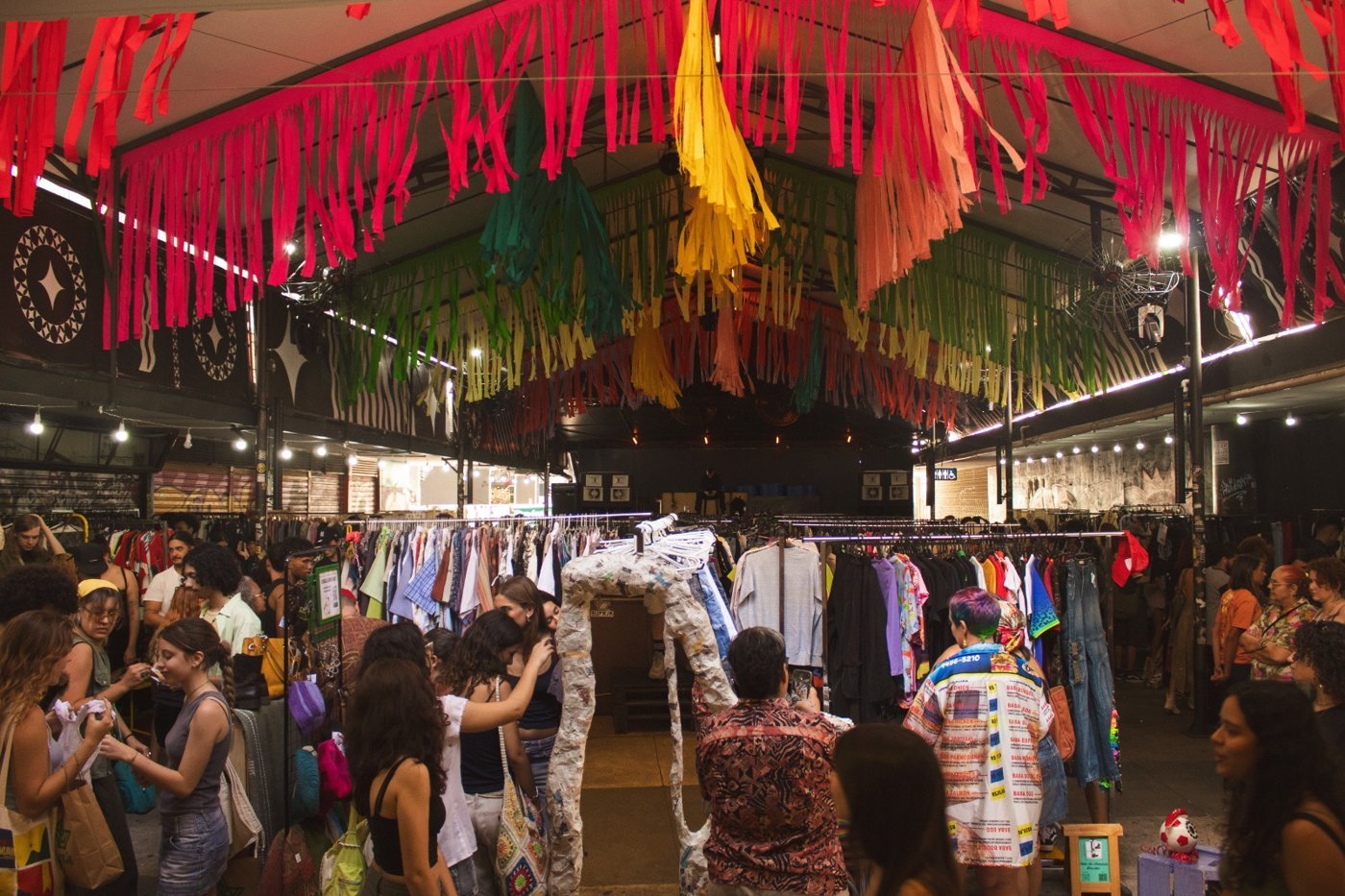 Araras de roupas sob bandeirolas coloridas e público circulando em edição anterior do Ao Desapego