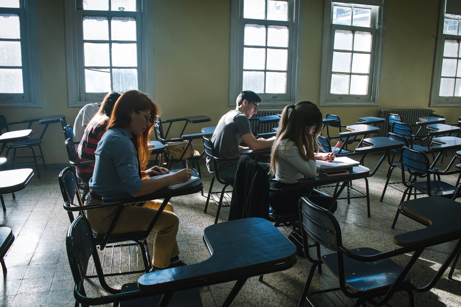 Estudantes sentados em carteiras universitárias escrevem em cadernos numa sala de aula iluminada pelas janelas