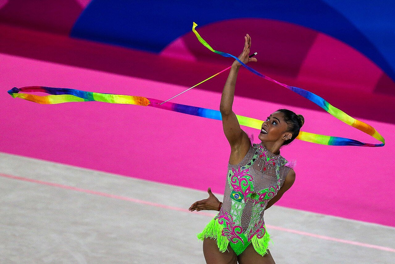 A ginasta Bárbara Domingos executa série com fita, de collant verde e rosa com a bandeira do Brasil