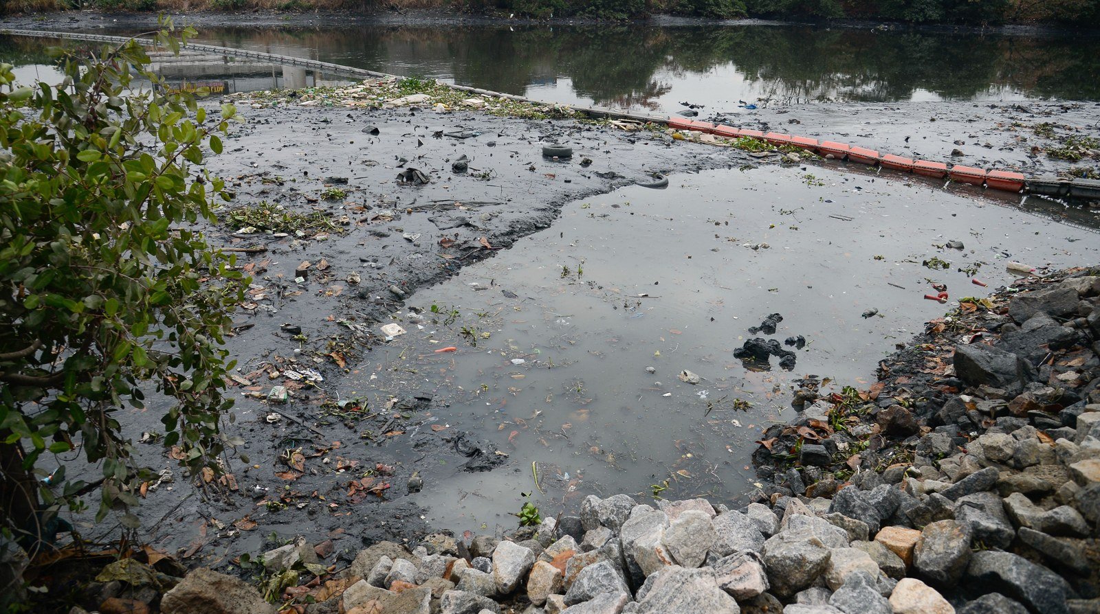 Ecobarreira instalada em rio da bacia da Baía de Guanabara retém resíduos flutuantes sobre a água escura