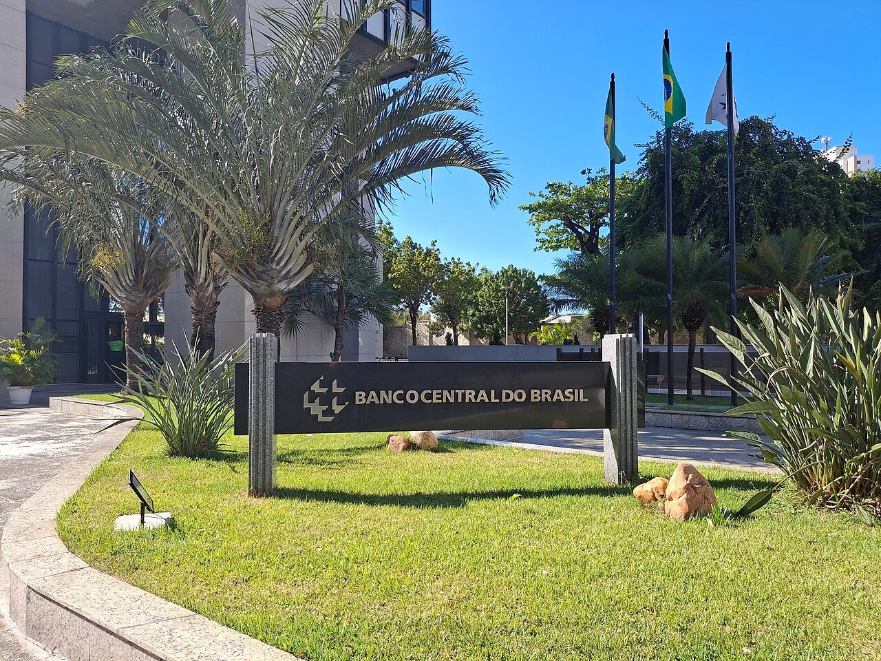 Placa com a identificação Banco Central do Brasil na entrada de um edifício, com bandeiras ao fundo