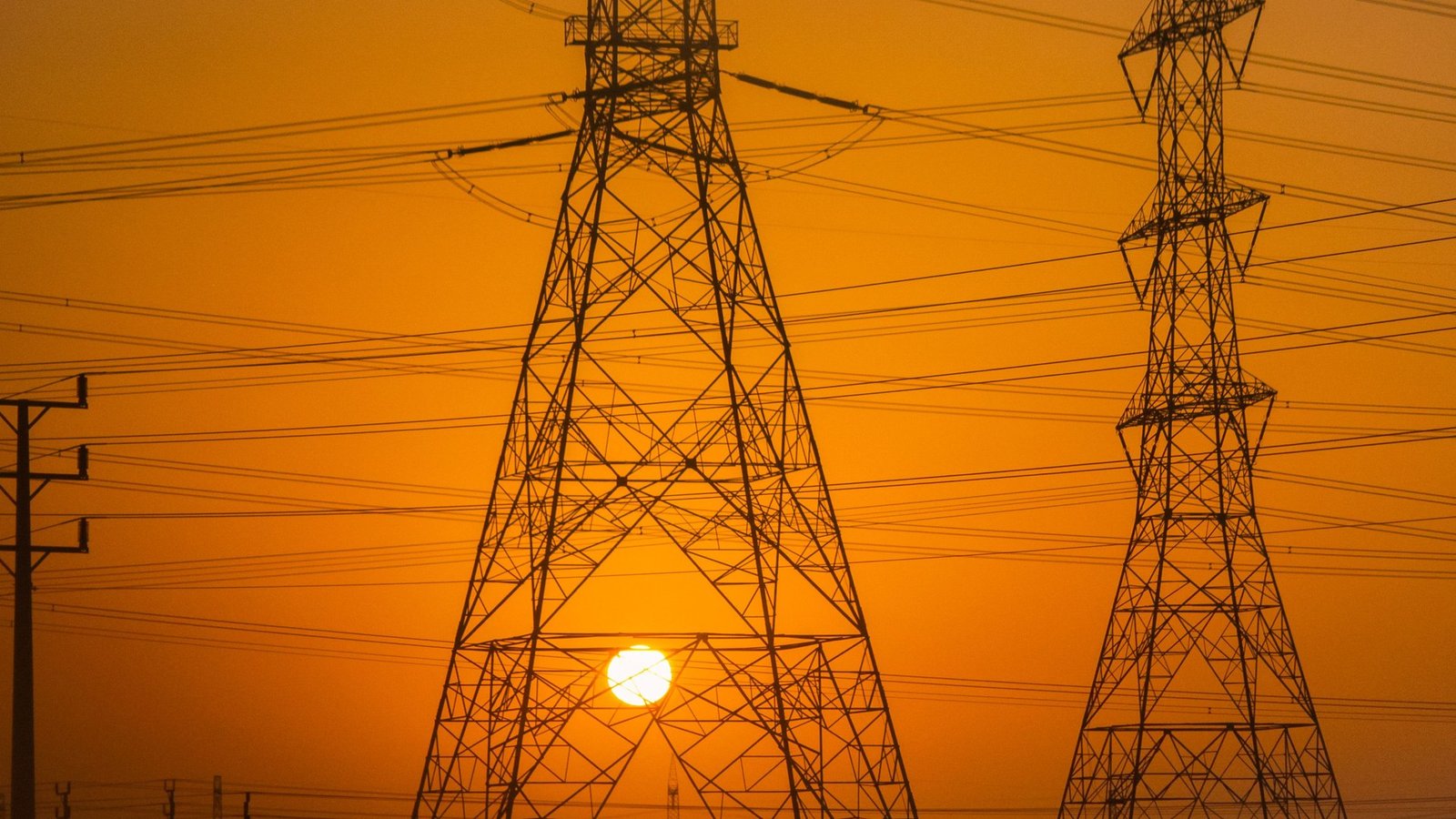 Silhueta de duas torres de transmissão de energia e vários cabos elétricos contra um céu alaranjado de fim de tarde, com o sol entre as estruturas