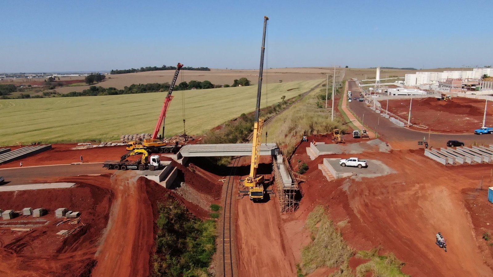 Vista aérea de canteiro de obras de ponte sobre linha férrea, com dois guindastes e vigas de concreto