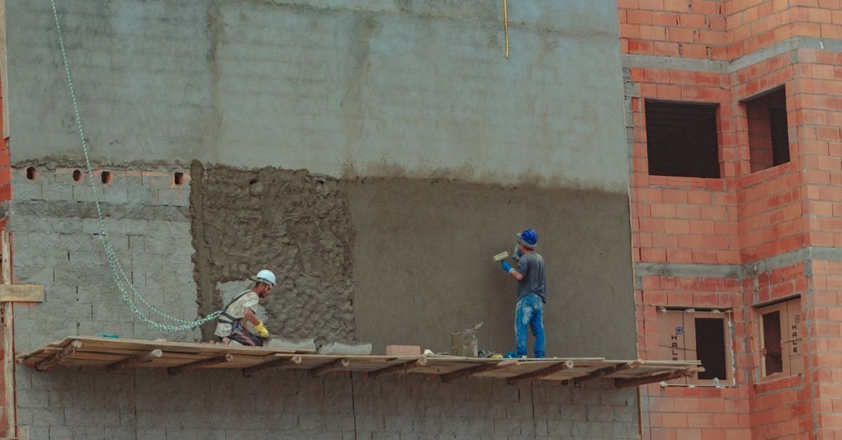 Dois pedreiros de capacete aplicam reboco na parede de uma construção de alvenaria inacabada