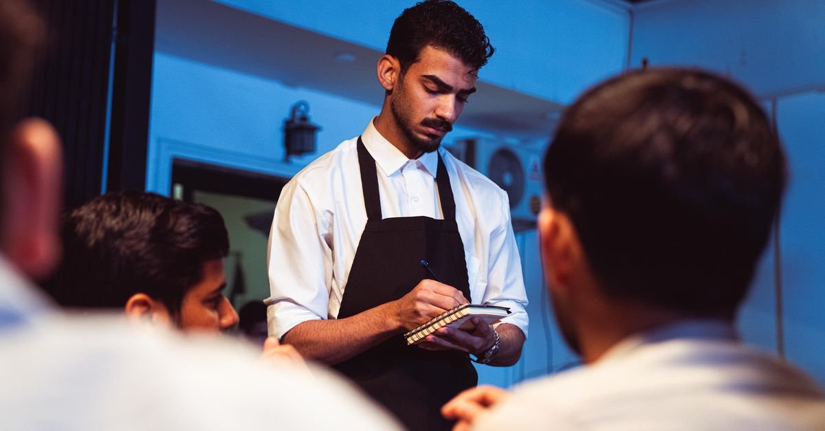 Garçom de camisa branca e avental preto anota o pedido de clientes em um restaurante