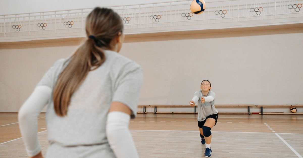 Jogadoras de volei durante treino em quadra coberta, uma delas se preparando para receber a bola