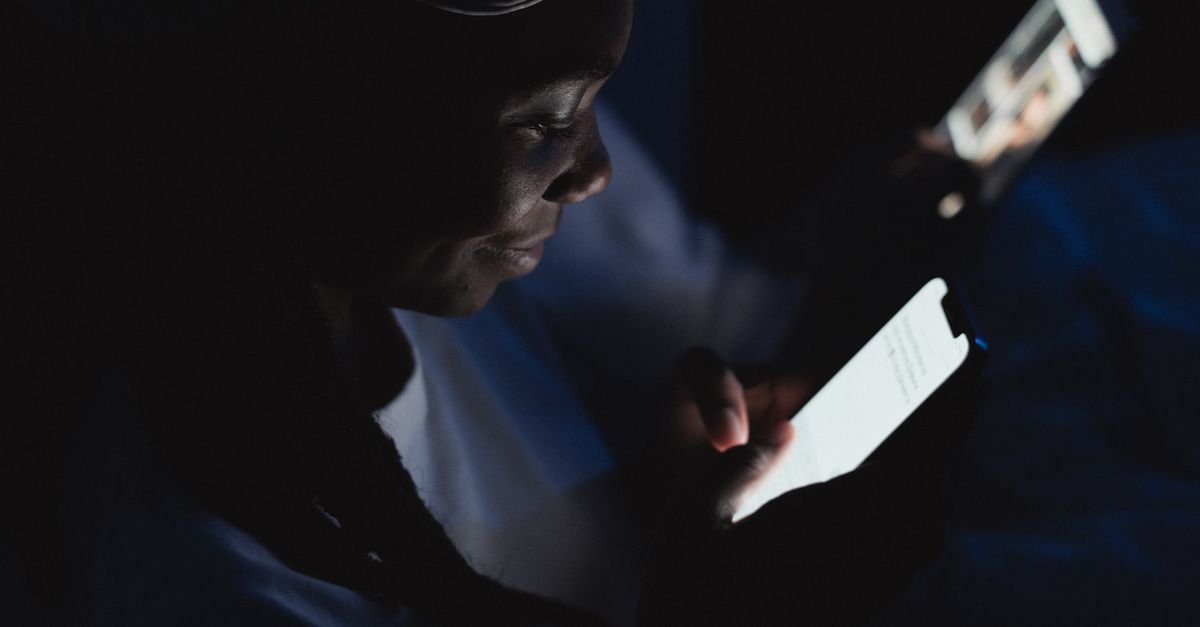 Pessoa usa smartphone em ambiente escuro, com o rosto iluminado pela tela do aparelho