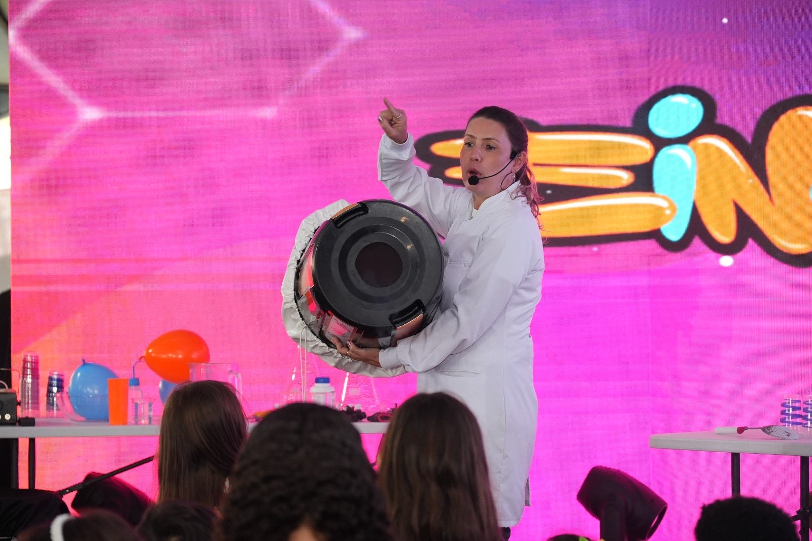 Apresentadora de jaleco branco faz experimento com canhão de ar durante show de ciência para plateia de crianças