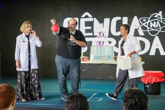 Einstein Jr. segura um balão vermelho durante show de ciência no palco do Ciência na Estrada, ao lado de duas apresentadoras de jaleco