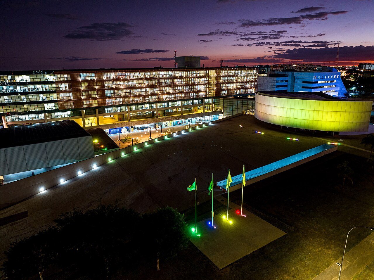 Sede da Câmara Legislativa do Distrito Federal ao entardecer, com o auditório circular iluminado e mastros com bandeiras em primeiro plano, foto de arquivo