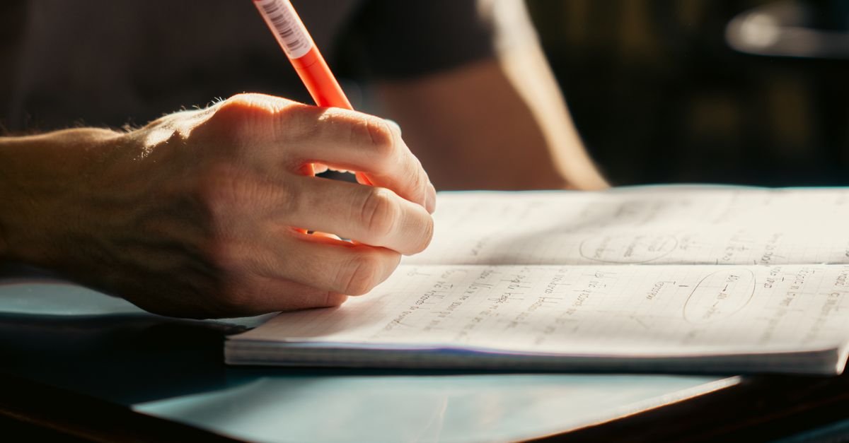 Mão de estudante escrevendo com lápis em caderno aberto sobre a mesa