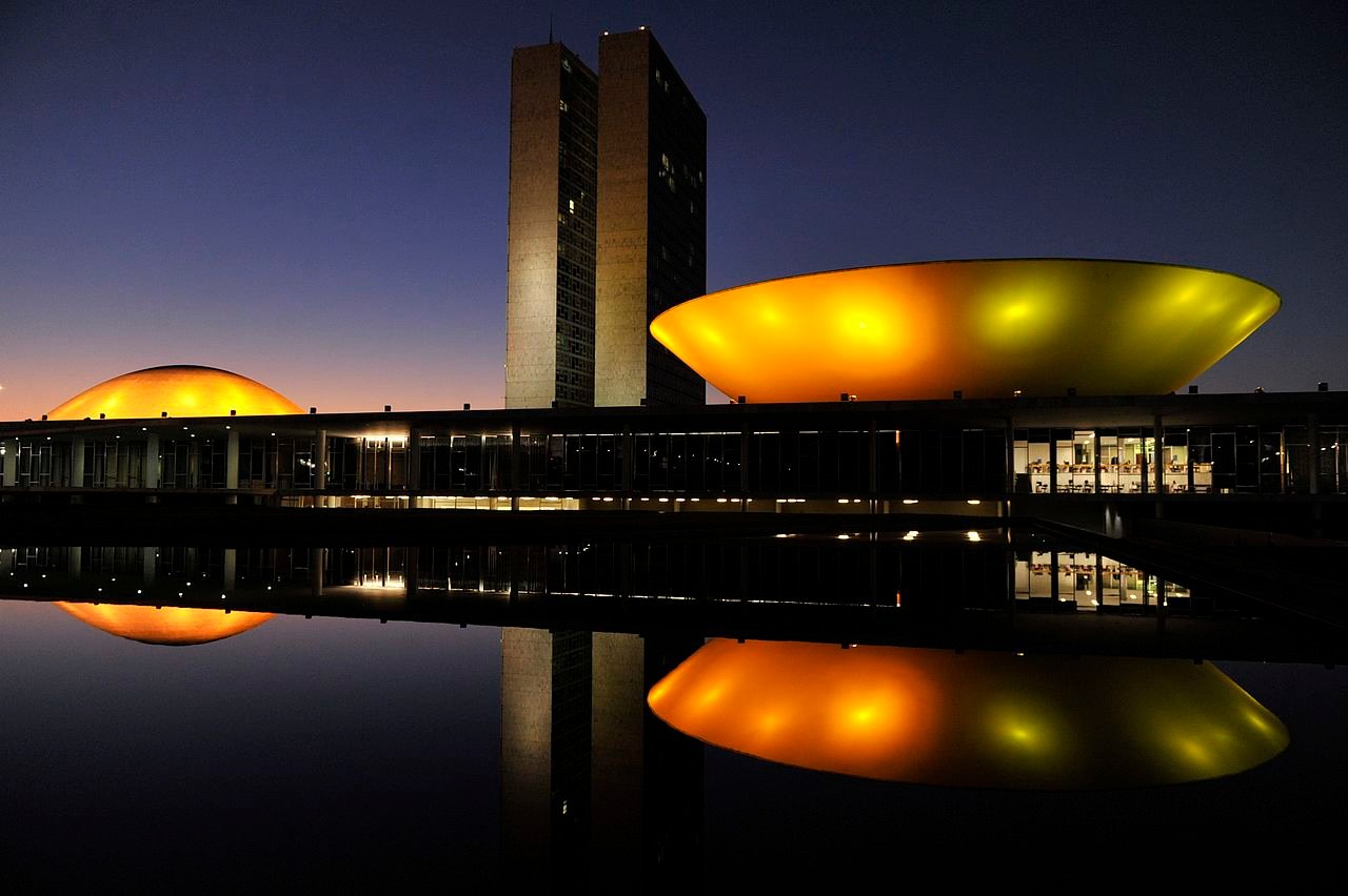 Congresso Nacional ao anoitecer, com as cúpulas do Senado e da Câmara iluminadas e refletidas no espelho d'água