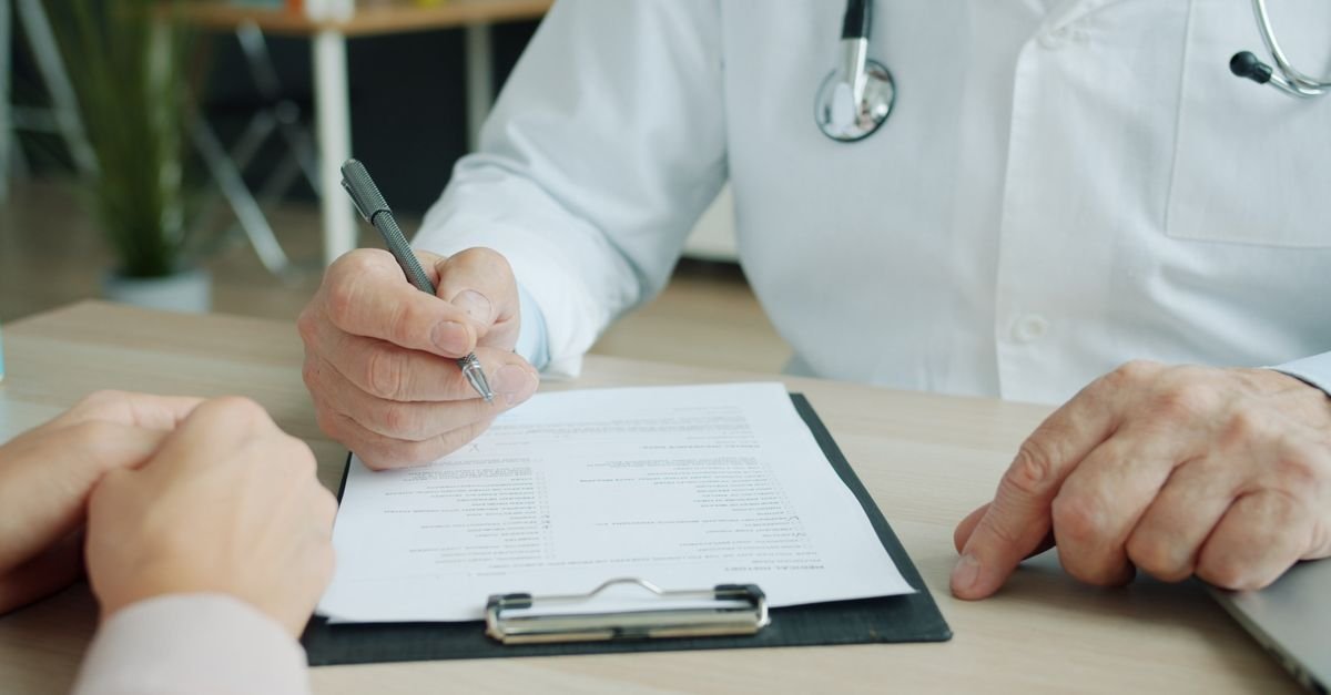 Médico de jaleco branco escreve em prancheta diante de paciente sentado à mesa