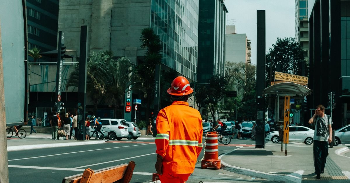 Trabalhador de uniforme laranja e capacete atravessa avenida movimentada