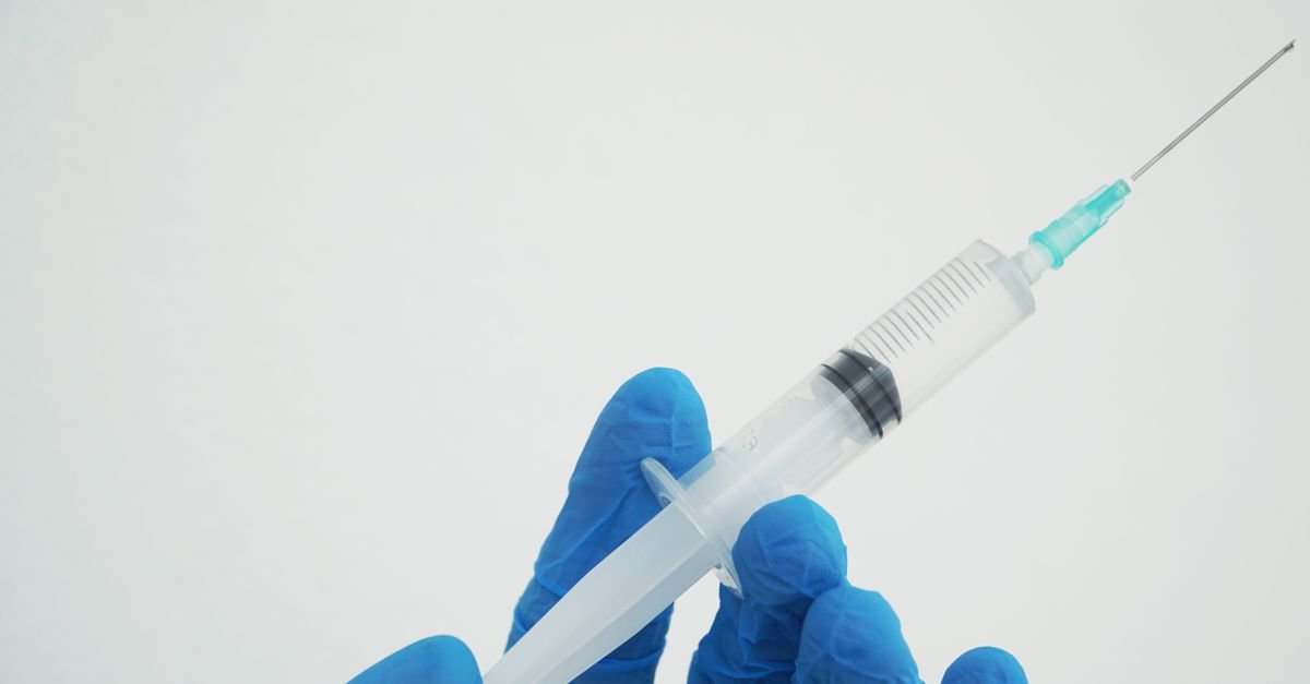 Mao com luva azul de procedimento segurando uma seringa contra fundo branco