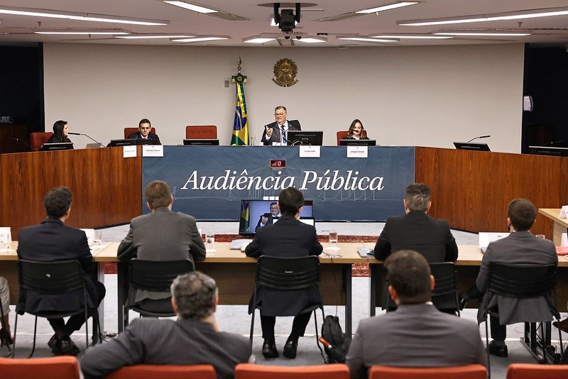 Mesa de autoridades e expositores sentados na sala de sessões da Primeira Turma do STF, com painel escrito Audiência Pública ao fundo