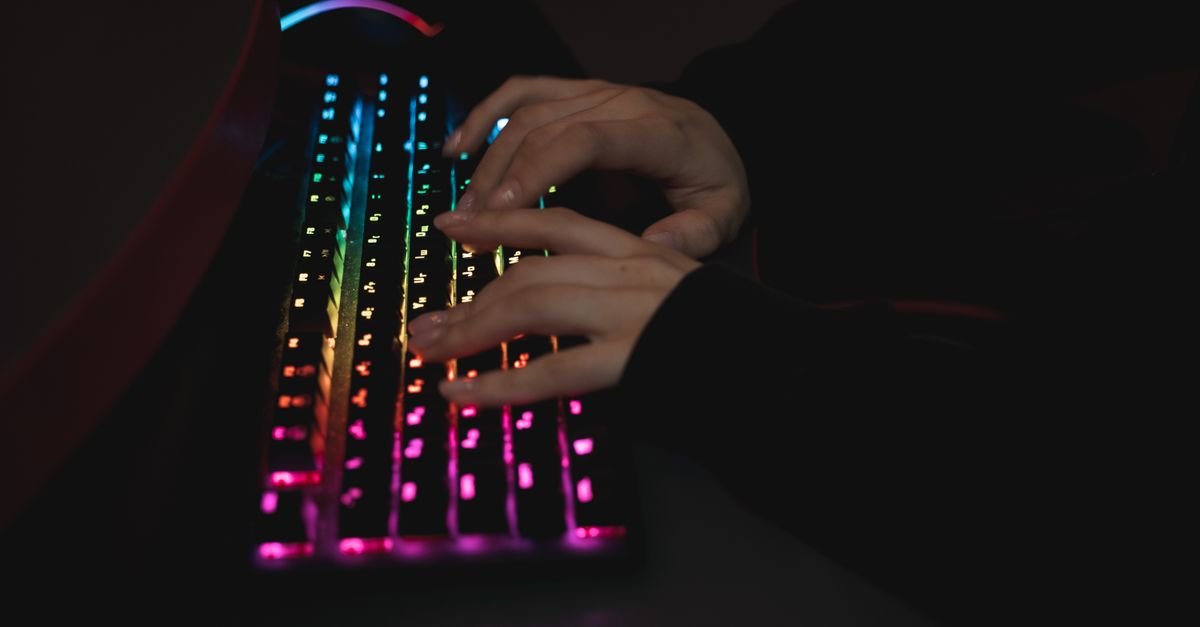 Mãos digitando em um teclado mecânico com iluminação colorida, em ambiente escuro