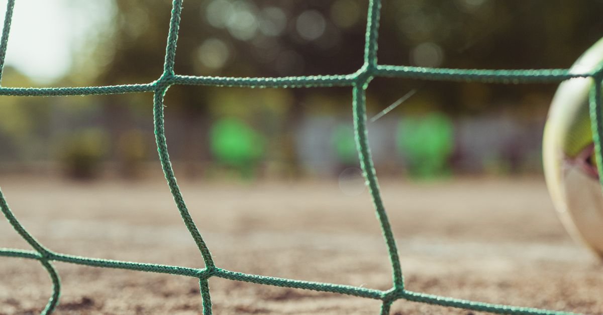 Ground level of green soccer net on rough terrain near ball on blurred background