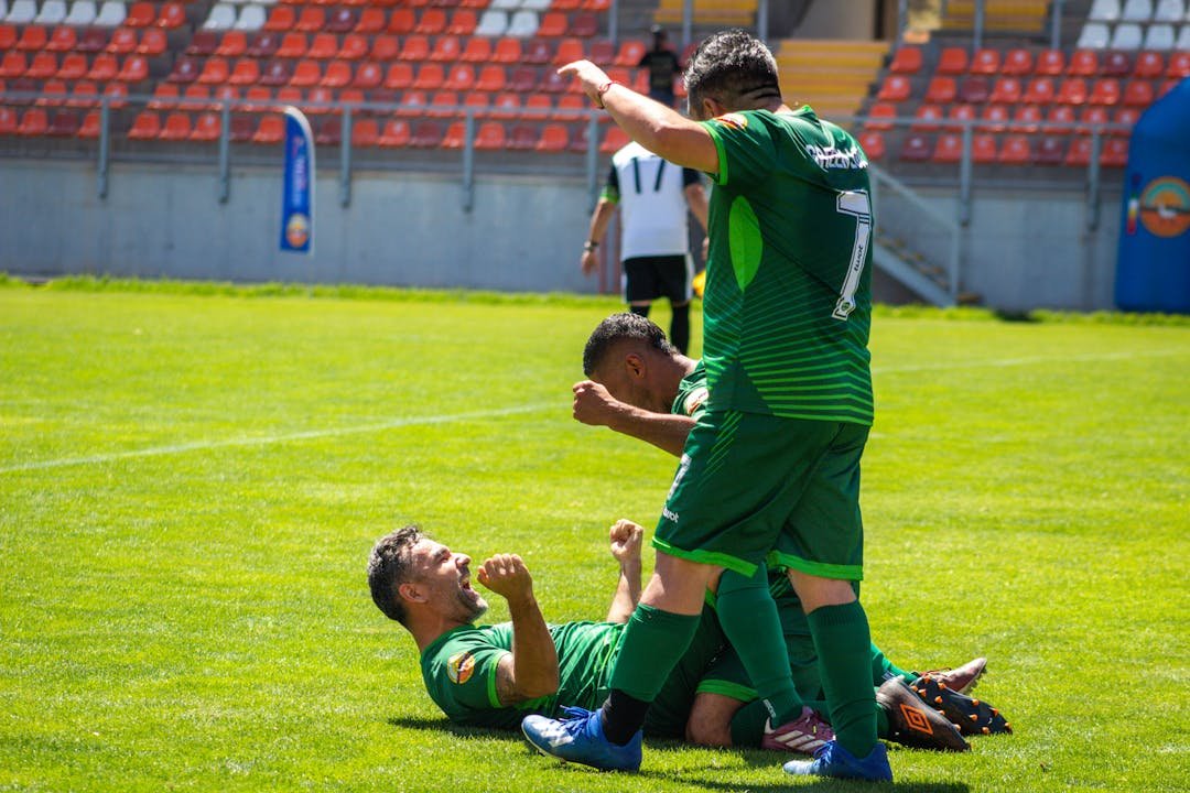 Jogadores de futebol comemorando gol em campo verde com arquibancadas vermelhas