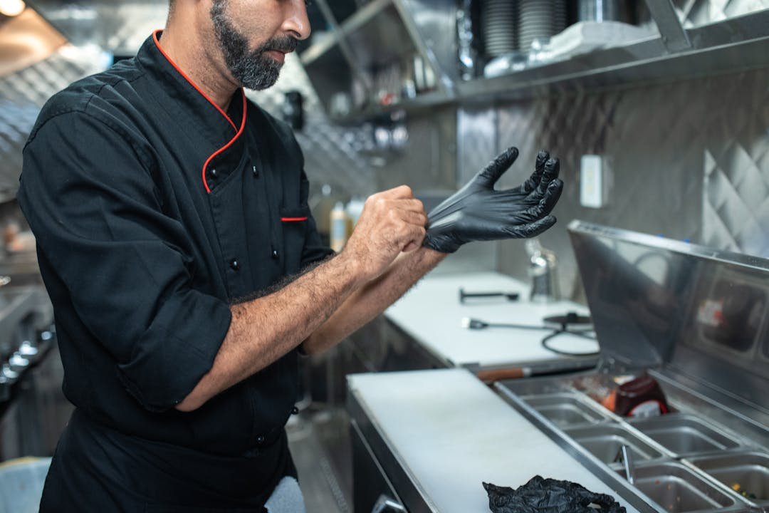 Profissional de cozinha com uniforme preto colocando luvas em ambiente de restaurante