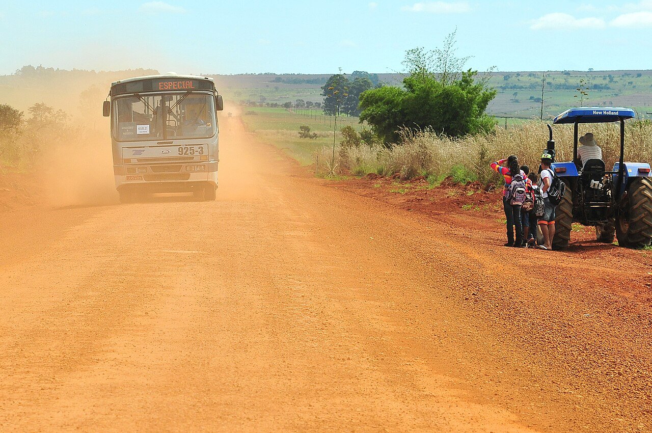 Ônibus público em estrada de terra com poeira e pessoas ao lado