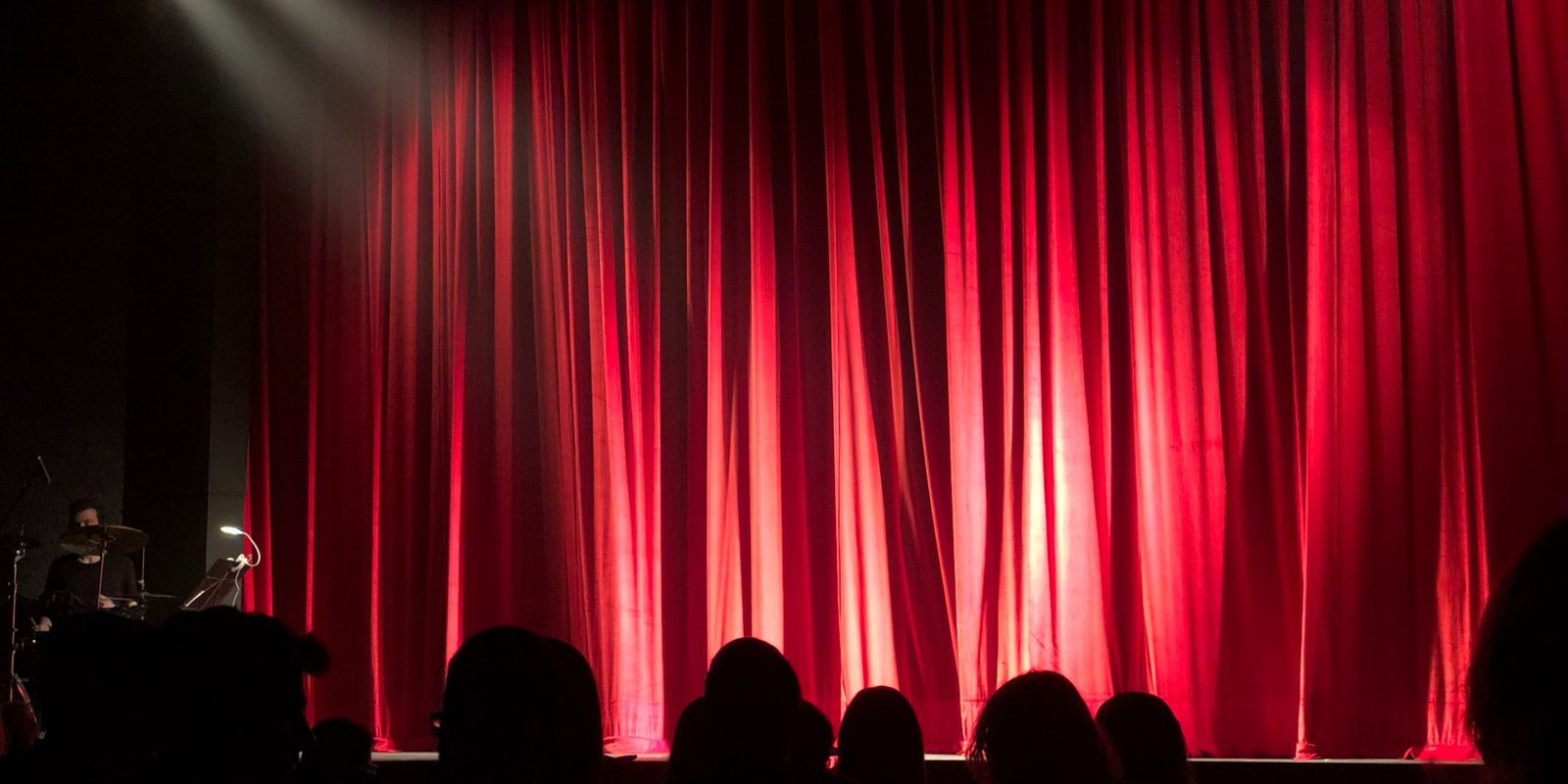 Cortina vermelha fechada em palco de teatro, iluminada por refletor, com plateia em silhueta
