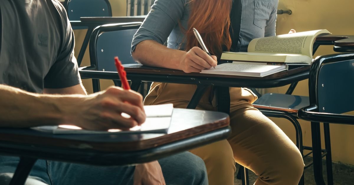Dois estudantes escrevem em cadernos apoiados em carteiras escolares durante aula