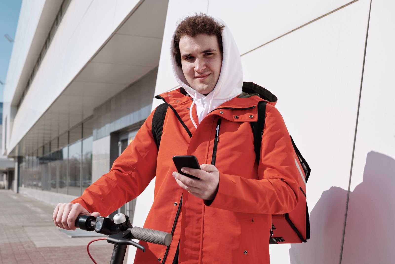 Entregador de jaqueta laranja com mochila térmica e celular na mão, ao lado de um patinete elétrico