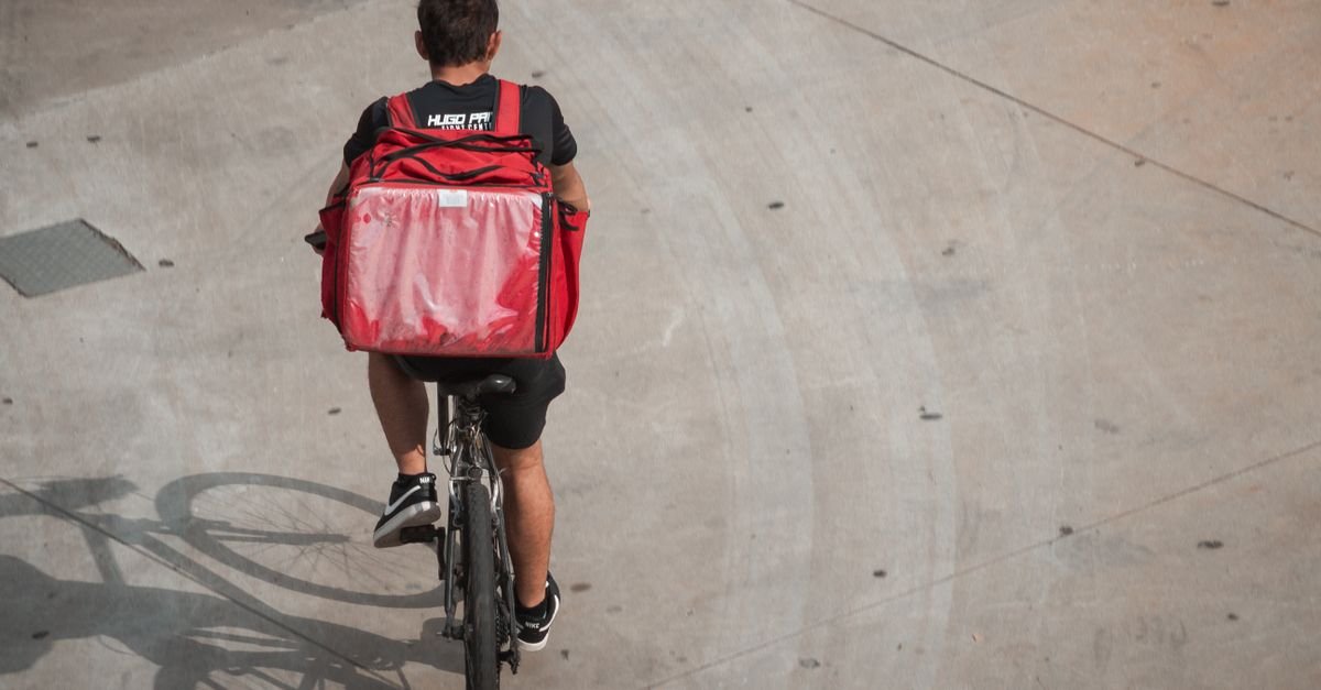 Entregador de aplicativo pedala com mochila térmica vermelha em via urbana
