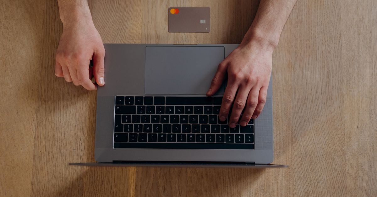 Mãos sobre o teclado de um notebook ao lado de um cartão de crédito em mesa de madeira