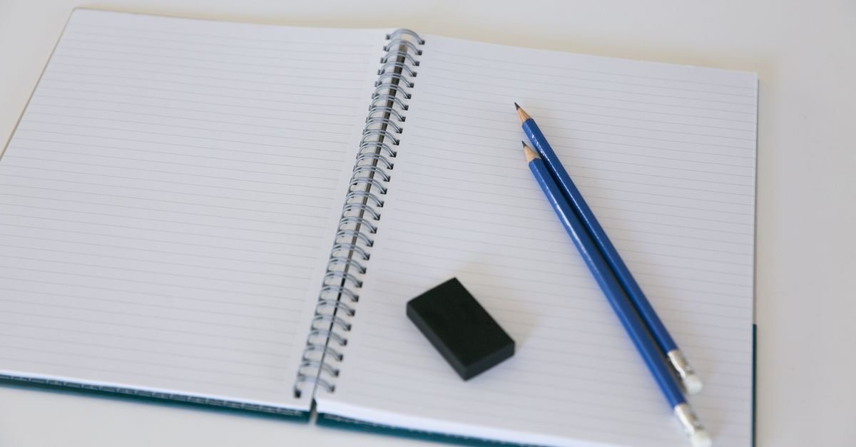 Caderno espiral aberto em folha pautada em branco, com dois lapis azuis e uma borracha preta ao lado