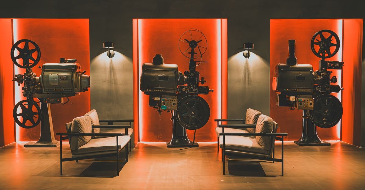 Três projetores de cinema antigos expostos em uma sala com iluminação vermelha e poltronas