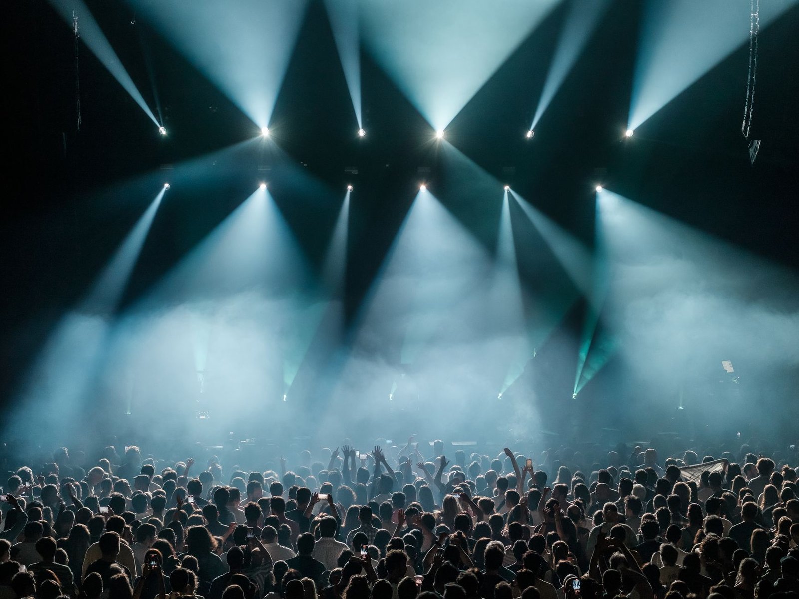 Plateia de um show em espaço fechado, de costas, diante de palco iluminado por feixes de luz azul e fumaça