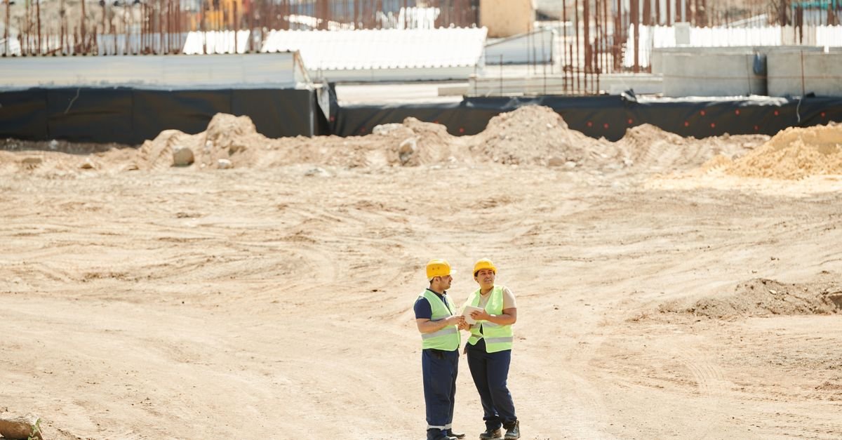 Dois trabalhadores com capacete e colete de segurança conversam em canteiro de obras