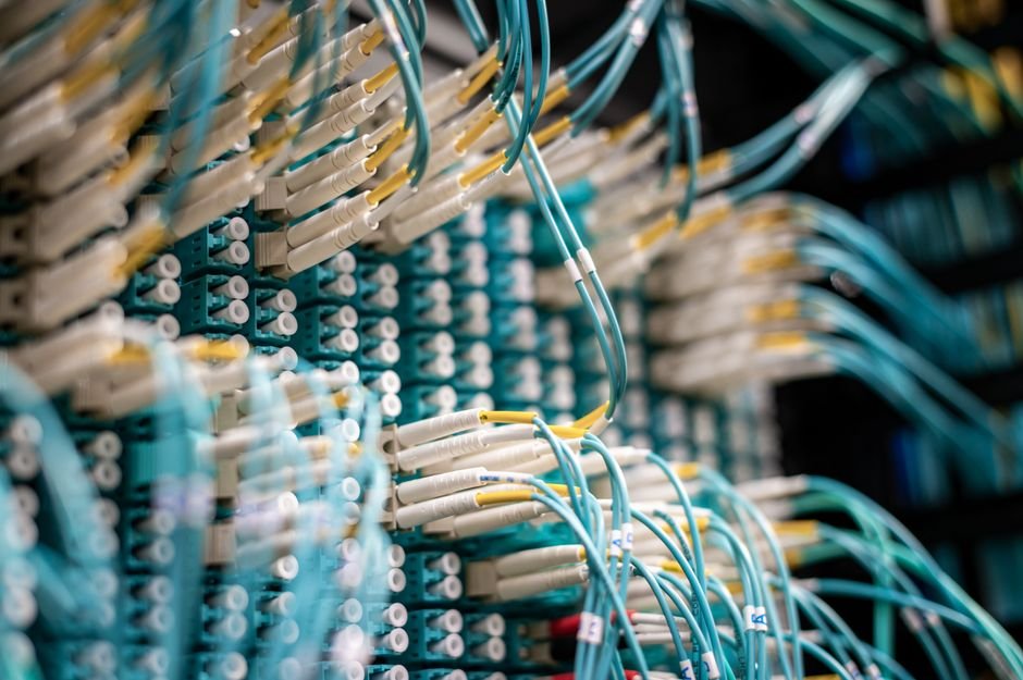Painel de fibra óptica com dezenas de conectores brancos e cabos azuis em um rack de telecomunicações