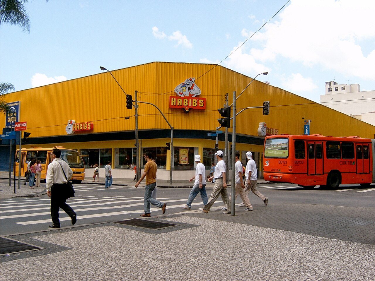 Fachada amarela de uma loja do Habib's em esquina de avenida, com pedestres atravessando na faixa