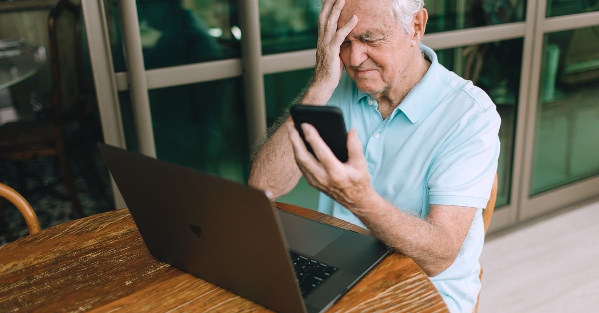 Homem idoso com expressão de preocupação segura celular diante de notebook