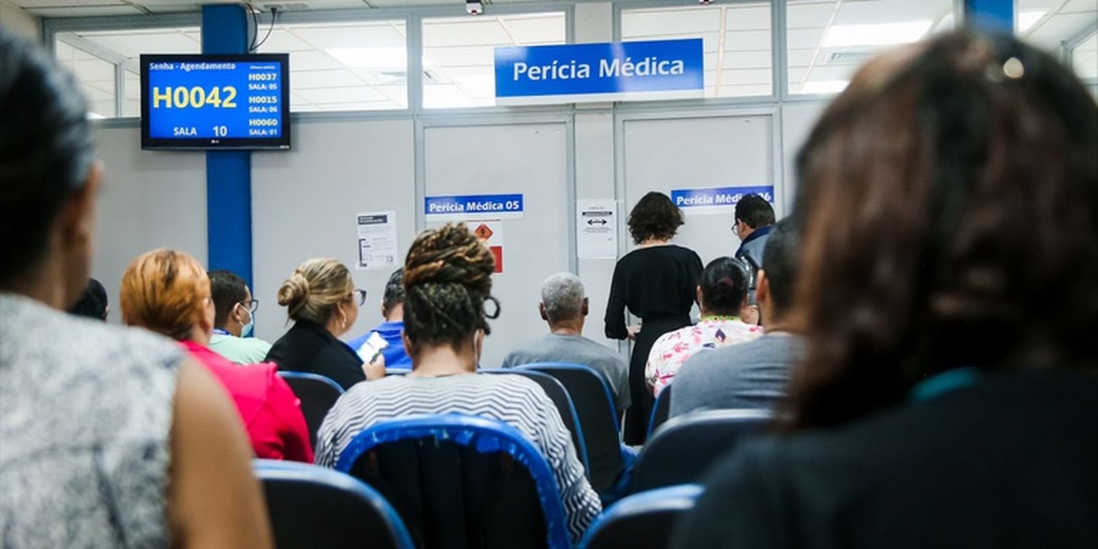 Pessoas sentadas em sala de espera de agencia do INSS diante de placa de pericia medica e painel de senhas