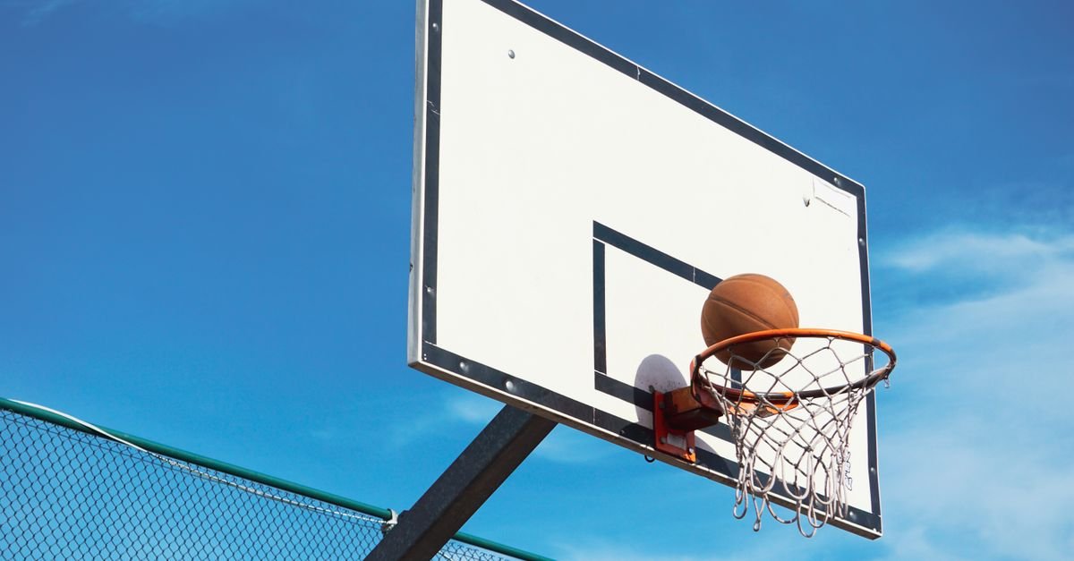 Bola de basquete entrando na cesta em quadra ao ar livre, com o céu azul ao fundo