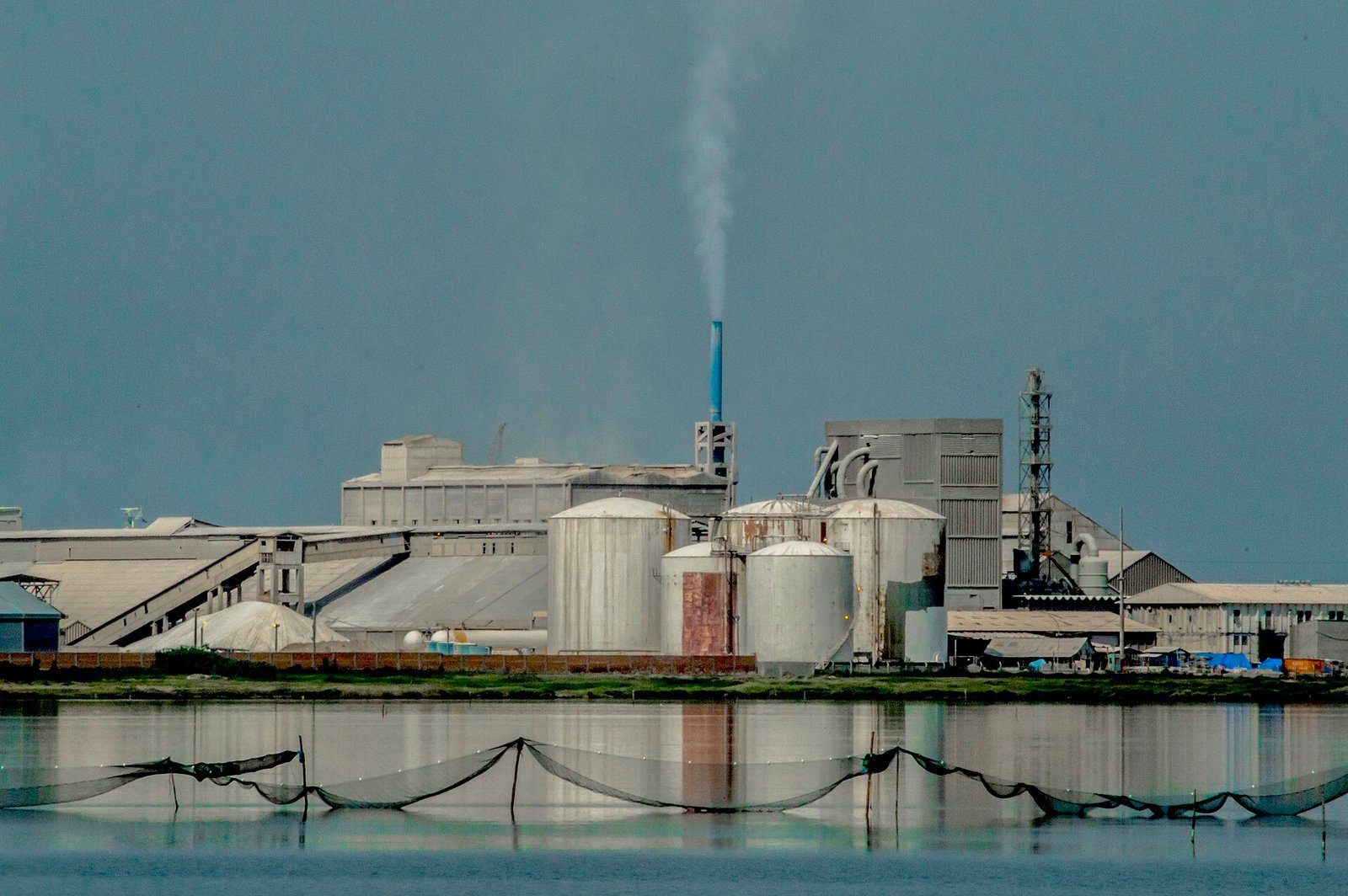 Complexo industrial visto de longe, com silos e chaminé, refletido na água