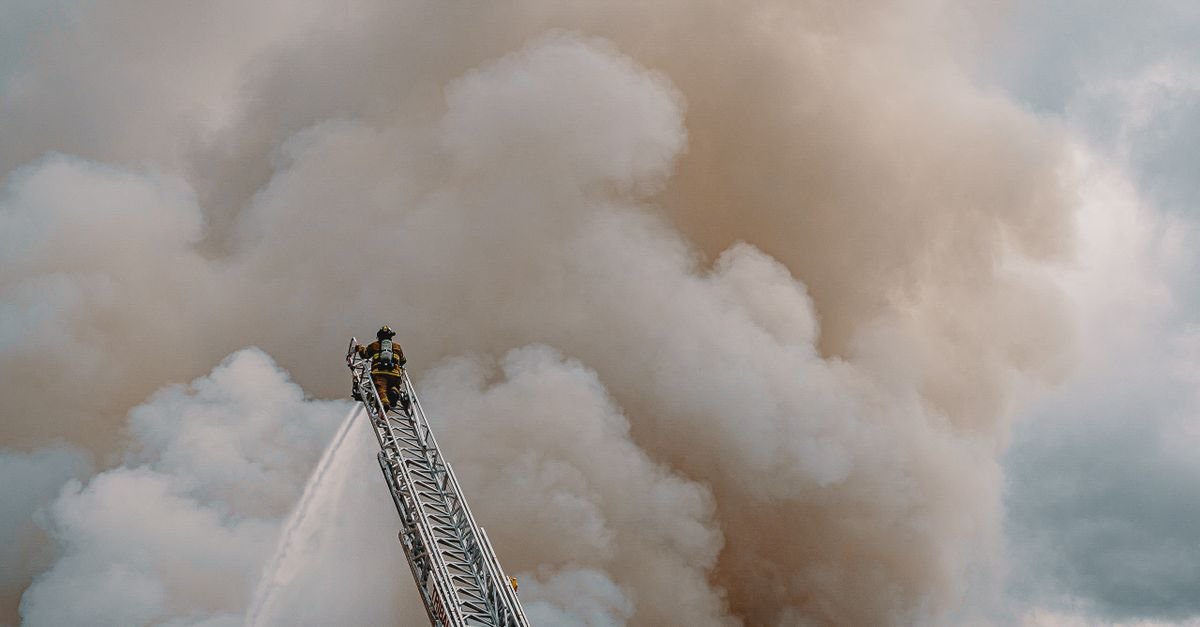 Bombeiro em escada aérea lança jato de água contra fumaça densa durante combate a incêndio