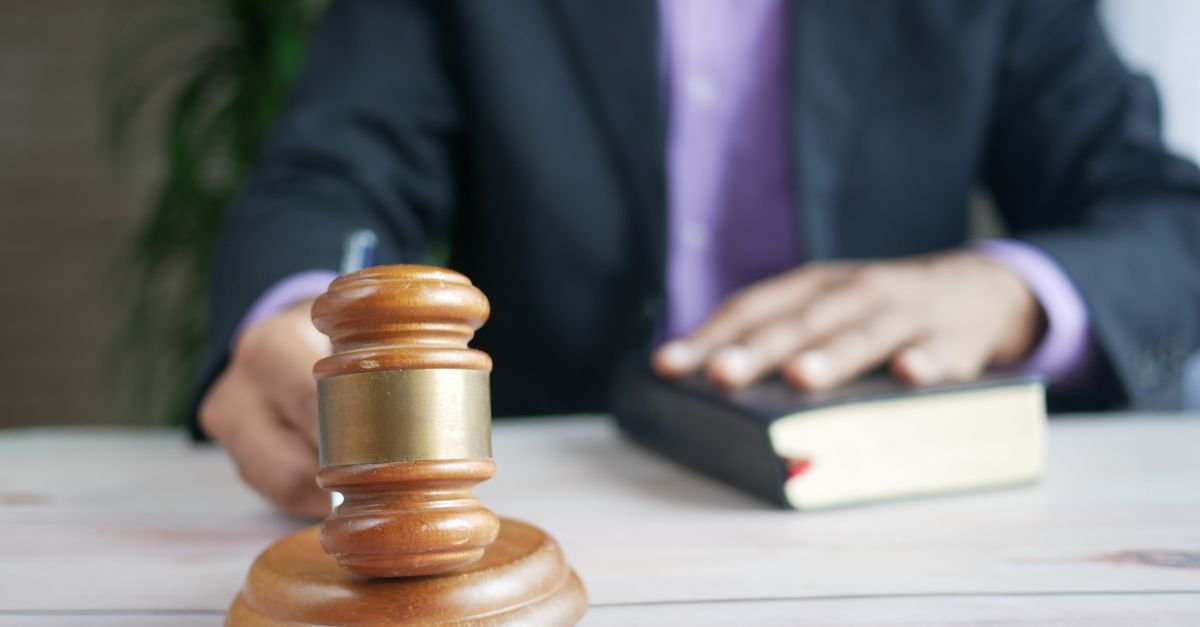 Martelo de juiz em primeiro plano sobre mesa, com homem de terno e livro jurídico ao fundo